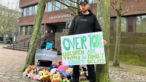 Simon Cobb has brown stubble and blue eyes. He is wearing a black hoodie and cap. Behind him is Portsmouth Family Court. He is standing next to a pile of plush toys and is holding a green and white placard.