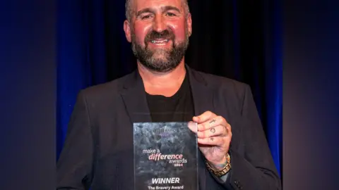 Steve Whitehouse is wearing a black suit and T-shirt underneath and is smiling holding his award and smiling in the photo. Steve has dark hair and a beard and is wearing a gold-coloured watch on his left hand and his fingers are also tattooed. 