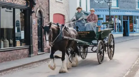 Ironbridge Gorge Museum Trust A two-wheeled carriage is being pulled by a large brown horse with two women in period dress sitting in it. There is a row of Victorian-style shops behind.
