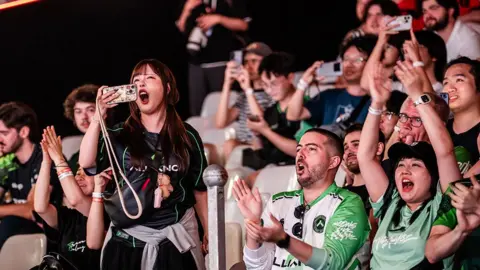 Esports Foundation A young female spectator wearing the black jersey of the Alliance esports team stands up from her seat, filming the action in front of her on her phone. She's yelling in support as she does so. Around her, other seated spectators are clapping or raising their arms and cheering.