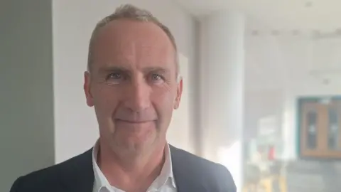 Neil Harrison stands in a university hallway, looking into camera. He is wearing a white shirt and dark suit jacket with no tie. He has receding grey hair and is smiling.