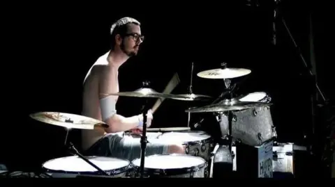 James Derrig A young man drumming. He is skinny and has an arm band around his right arm