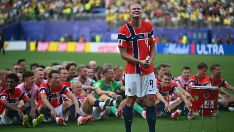Getty Images Erling Haaland standing on a football pitch, about to bang a drum as his teammates do the row on the pitch behind him. 
