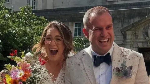 Kim and Gareth Gladman grin as they are showered with confetti on leaving their wedding ceremony at Leeds Civic Hall. They are in their cream wedding outfits and she is holding a floral bouquet.