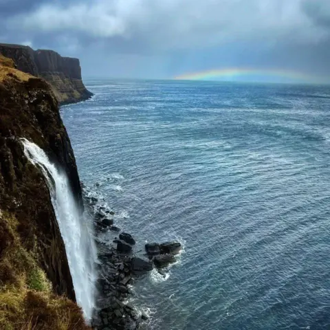 Peter Xu A distant rainbow over the sea, with a waterfall going into the water in the foreground.