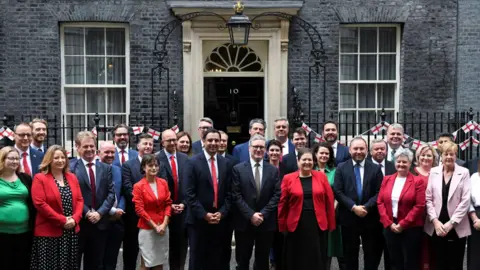 Reuters Scottish Labour MPs standing in front of 10 Downing Street. Sir Keir Starmer and Anas Sarwar are standing in front on the group in dark suits. The door to number 10 is behind them and is black.