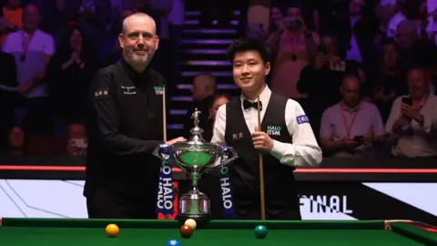 Getty Images Mark Williams and Zhao Xintong pose for a photo prior to a World Snooker Championship match at Crucible Theatre. They both hold a large silver trophy, resting on the snooker table in front of them, and a large audience behind them. 