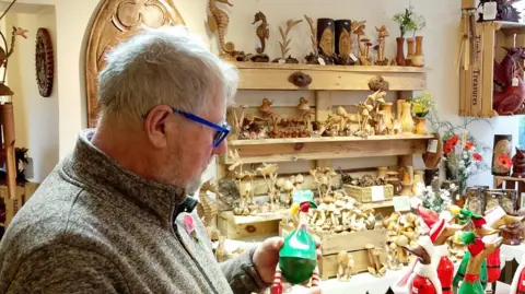 David in his shop surrounded by wooden items, some of which are painted in bright green and red, with some yellow. He is holding a wooden painted duck.
