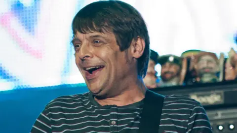 PA Media Mani on stage in a green T-shirt with light green horizontal stripes. He has short brown hair.