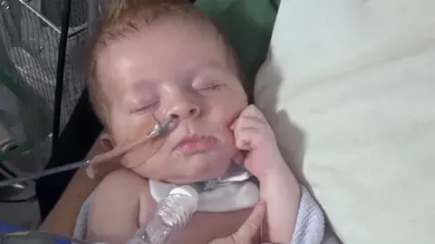 Family Baby Joshua is seen sleeping with his eyes closed, lying in his parent's arms. Joshua has light hair which is spiked at the top of his head. He has a tube into his nostril, secured with a nude plaster. He also has a white tube, secured with a white bandage onto his neck. 