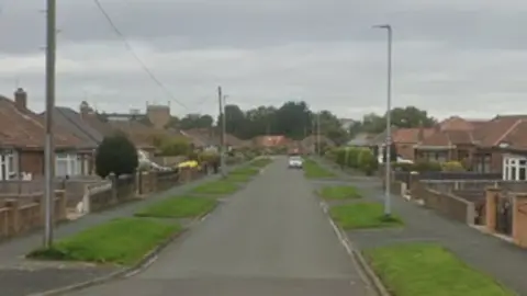 The image shows a quiet residential street in a suburban area. The viewpoint is positioned in the centre of the road, facing straight ahead.