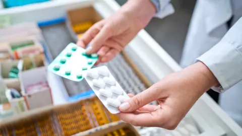 Getty Images Hands holding two blisters of tablets over a counter with a range of products in it