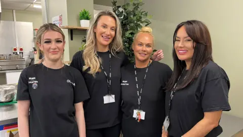 A teenage girl with blonde hair, two women with blonde hair and a woman with brown hair are standing together in a café kitchen.
They are all wearing black short sleeved t-shirts.