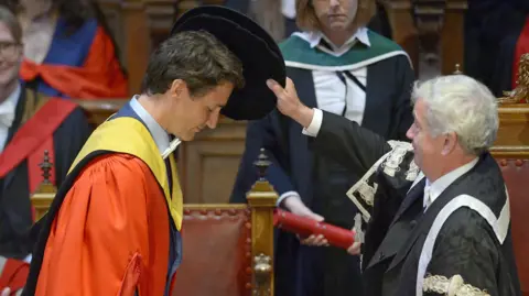 A person in a red academic gown bows during a university degree ceremony as an official in ceremonial robes lowers a black academic cap over the person's head, while attendees and faculty members look on in a wood-panelled hall.