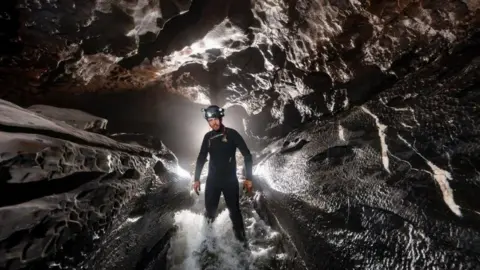 Mark Burkey George Linnane is photographed underground in a cave. He is stood the centre of the image, standing in water. He is wearing a wet suit and a satfey helmet.