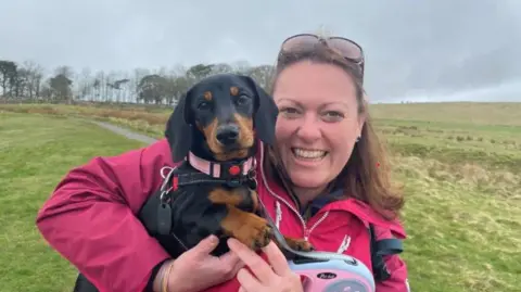 Kate Harrison is a young woman holding dachshund Bobbi in her arms. She is wearing a pink anorak and behind her is a long stretch of hillside with a path and a row of trees at the top 