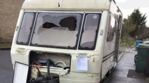 An old caravan with smashed windows and the front cover of the gas locker missing. There is a notice on the front of the caravan and the side entrance door is open. 