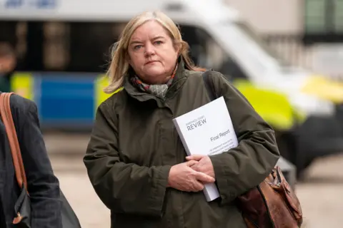 PA Media Baroness Casey clutches a copy of her report into the behaviour and culture at the Metropolitan Police Service. She is walking towards the camera wearing a green, waxed jacket and is carrying a leather shoulder bag.