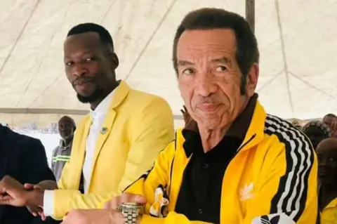 O inocente Selatlhwa Ian Khama em agasalho amarelo aponta para seu relógio.