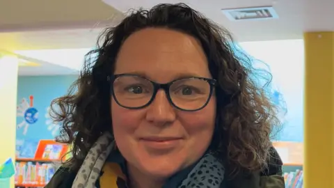 Jacob Ottaway/BBC Laura John smiles at the camera. She wears black glasses and has short brown curly hair. She wears a scarf around her neck and a coat. 