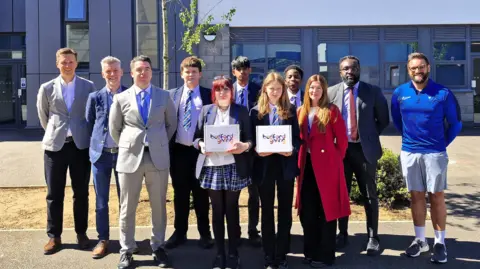 Bedford Giving A group of people standing outside a school. Two people are holding up certificates. There is a tree behind them. 