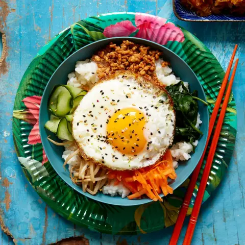 Future Publishing via Getty Images Bibimbap, a Korean dish featuring rice and various vegetables such as carrots, spinach, bean sprouts and a fried egg on top, served with chopsticks, and a side dish of kimchi.