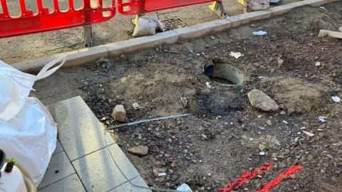 A close up of a pavement that has only partially been covered in paving slabs, with a set of red plastic railings guarding it.