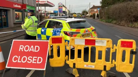 BBC A road closure sign, police car and a police officer standing near some police tape at the foreground of the image. In the background, the A38 stretches out, with an ESSO garage on the left and some more police vehicles in the background.
