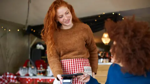 Getty Images Cliente em um café fazendo pagamento sem contato com celular para uma garçonete vestindo um avental xadrez vermelho e branco