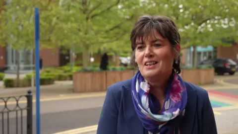 A woman stands in front of a road with greenery behind her in a town centre. She is wearing a navy blue jacket and blue pink and white patterned scarf.