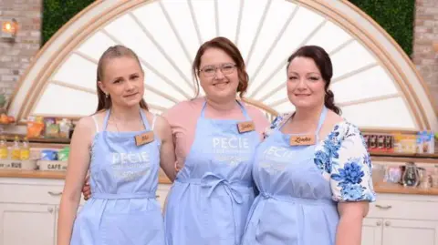 Three women stand side by side in a bright kitchen set, each wearing a light blue apron with a name tag. Behind them are white cabinets, baking equipment, and a decorative arch with a sunburst design.