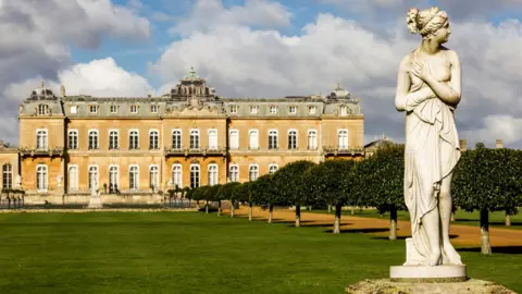 A classical white marble statue stands in the foreground of a formal garden, with neatly trimmed trees lining a path. Behind it, a large historic manor house with many windows stretches across the background under a partly cloudy sky.