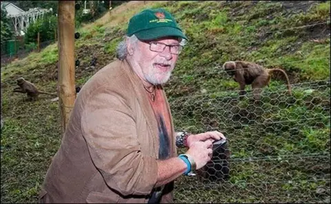 A man with grey hair and glasses with a green baseball cap and brown jacket. He is standing by an enclosure bounded by chicken wire, with a small primate on the grass behind. 