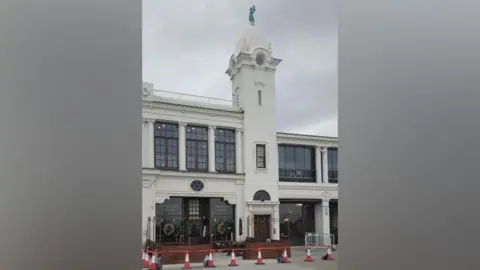 The Split Chimp which has a dark brown wood shop front cordoned off by orange and white cones. The shop is part of the white Spanish City building. The green dancing lady statue can be seen directly above the bar on top of a domed tower.