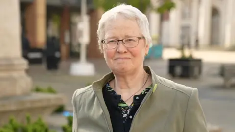 Chrissie Brown wears has short white hair and glasses. She wears a green jacket and black blouse.