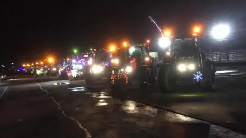 Several tractors are parked alongside one another, in the dark, on a large tarmac area. They have headlights on full and multiple Christmas lights as decoration. 