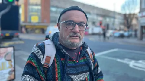 An older man with a grey beard, glasses and a black fisher mans hat. He has a multi coloured pattern hooded top and is wearing a grey bag pack. A city street can be seen behind him out of focus.