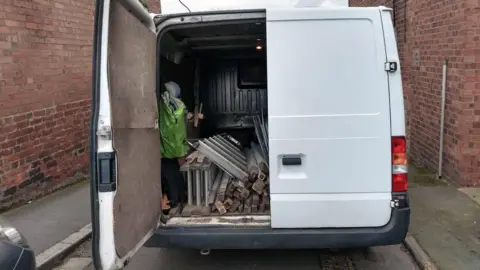 The back of a white van with one of the rear doors open, showing the tools it is loaded with