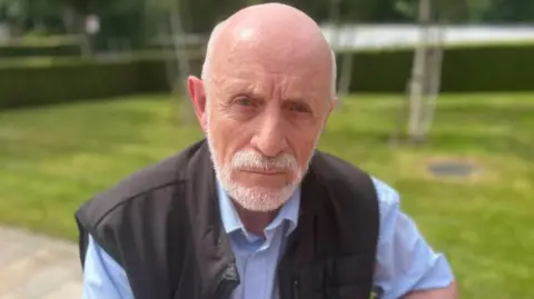 A man with a white beard looks into the camera. He is bald and is wearing a blue shirt and black gilet jacket.