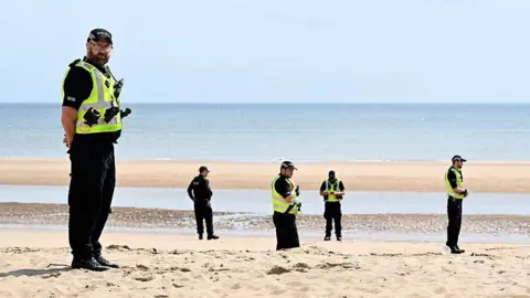 Getty Images Seis policiais do sexo masculino, vestindo uniformes pretos e coletes de alta visita em uma praia de areia com o mar ao fundo