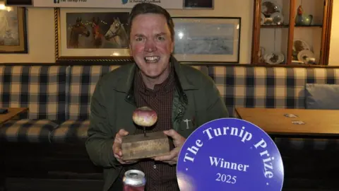 Trevor Prideaux Ali Can holds a block of wood with a turnip impaled on a nail and smiles at the camera. He is in a pub and wears a red shirt and green coat. There is a blue plaque with 'The turnip prize winner 2025' written on it.