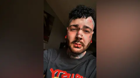 Harry Jackson - a man with a dark, curly, mohawk hairstyle. He also has several facial piercings and tattoos, as well as tattoos on his neck and head. He is looking at the camera and the room behind him is dark.