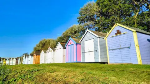 Weather Watchers/MrBlueSky Colourful beach huts at Felixstowe lined up on a grass area.