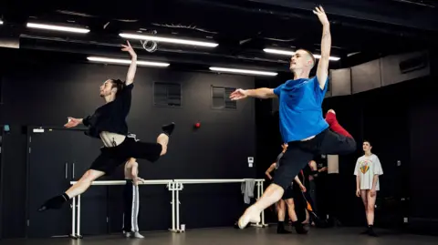 In a black dance studio there are two male ballet dancers mid-air in a pose with stretched out right legs and left legs curled behind them. Their left hands are stretched high and their right hands are stretched to their sides. There is a woman in a long T-shirt standing watching them. In the background is a barre.