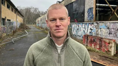 Paul Oster has a short buzz cut with light hair. He is wearing a light green zipped jacket. He is standing. Behind him is the derelict site.