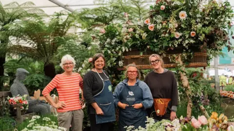 Four women standing in a line and smiling. Three are wearing glassed and two have blue aprons on. They are stood in front of a funeral flowers display which has flowers bursting out of an elevated coffin next to the a sculpture of a man sat on a bench.