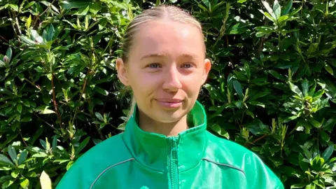 A close-up of Amie Donaghey. She is a young woman with slicked back, long blonde hair. She is wearing a green zip jacket and she is stood in front of a large bush.