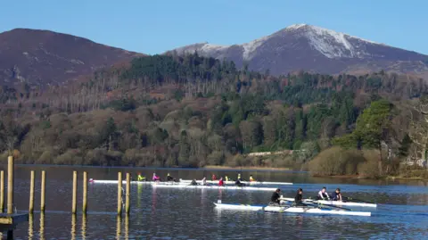 Multiple boats on a lake being rowed by people. The lake is clear and there is a mountain and trees in the background. 