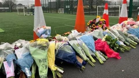Floral tributes fringe a sports field which has both football goals and rugby posts. Behind the row of flowers are several orange cones 