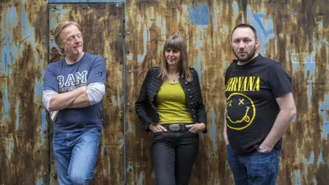 Two men and a woman are standing in front of a wall that is metal and rusted. The man on the left has short blonde hair and a white goatee. The woman has brown shoulder length hair. The man on the right has short black hair with black facial hair. He is wearing a black and yellow Nirvana t-shirt